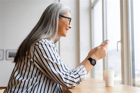 Smiling White Haired Mature Woman Drinking Coffee And Using Cellphone