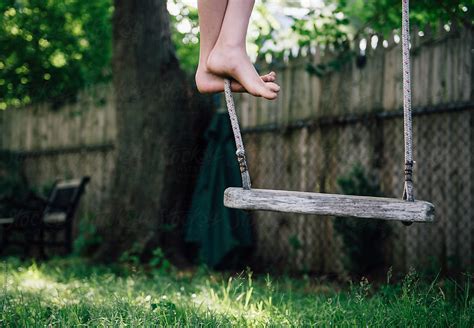 Child With Bare Feet Stands On A Wooden Swing In His Back Yard By