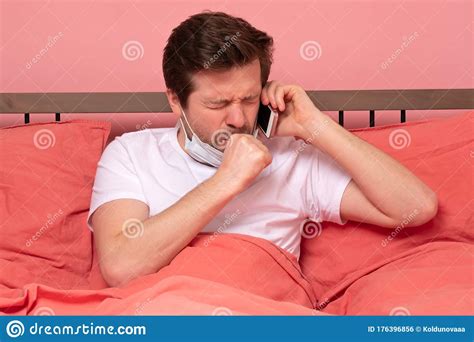 Sick Young Cauacsian Man Coughing While Lying In Bed And Speaking On