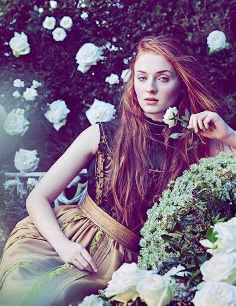 A Woman With Long Red Hair Sitting In Front Of White Roses And Greenery Holding A Flower