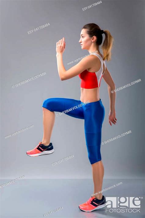 Young Energetic Woman Walks Highly Lifting Knees And Waving The Arms In The Gym Over Grey