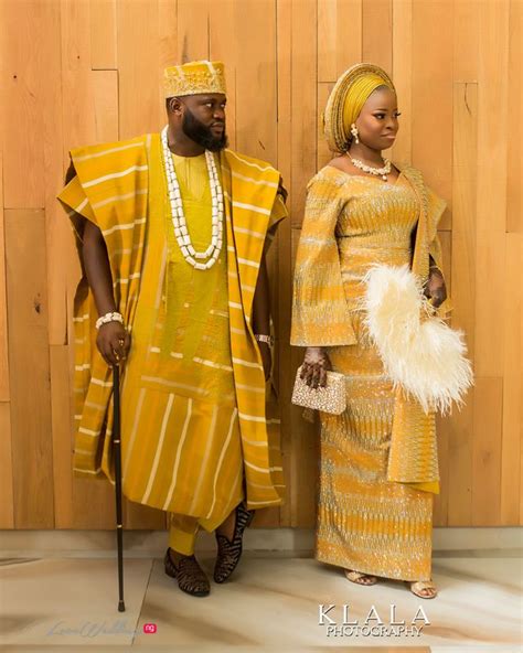 Two People Standing Next To Each Other In Front Of A Wooden Wall Wearing African Clothing