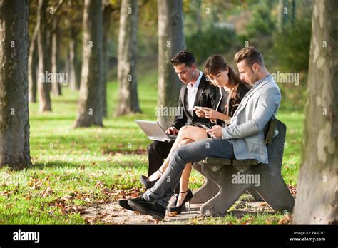 Three Business People Sitting On A Park Bench Using Laptop Stock Photo