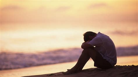Sad Depression Man Is Sitting Alone On Beach Sand In Blur Sky