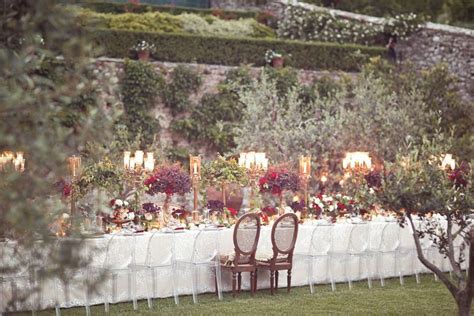 An Outdoor Dinner Table Set Up With White Linens And Chairs Surrounded By Greenery