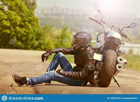 Biker In Outfit Sits Next To Motorcycle On Asphalt Motorcyclist Rests