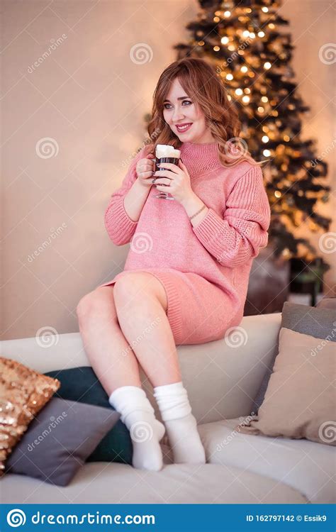 Beautiful Girl In A Pink Sweater And Socks Sits On A Sofa With A Cup Of Hot Drink In Her Hands