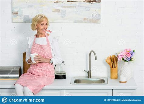 Attractive Senior Woman Sitting On Kitchen Counter With Cup Of Coffee