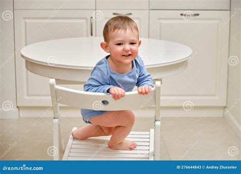 Toddler Baby Climbs Onto A Chair At The Kitchen White Table A Child In