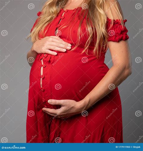 closeup pregnant woman in red dress holding her belly while standing over gray wall background