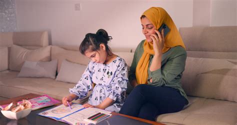 أم عربية في المنزل ، تقوم بإجراء مكالمة هاتفية بإستخدام الهاتف المحمول، تجلس إبنتها بجانبها تقوم