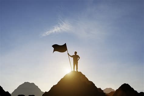 Silhouette Of Businessman Holding Flag On The Top Of Mountain With Over