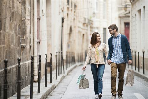 A Couple Walking Along A Narrow City Street With Shopping Bags Stock