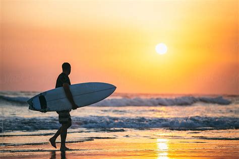 Man Carrying Surfboard Along Beach At Sunset Del Colaborador De