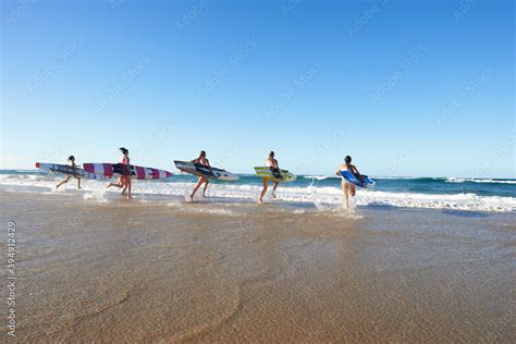 Back View Of Team Of Female Surf Lifeguards Training And Running Into Sea Holding Ocean Surf