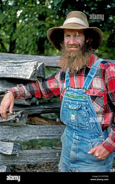 A Bearded Mountain Man In Plaid Shirt And Denim Overalls Relaxes By A