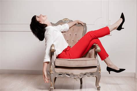 흰색 셔츠와 빨간 바지에 어두운 머리를 가진 매력적인 젊은 여성의 스튜디오 초상화는 일반 흰색 배경에 고전적인 의자에 앉아 보시