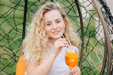 Outdoor Shot Of Pleasant Looking Female With Curly Light Hair Drinks Fresh Cold Drink Rests In