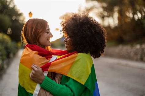 multiracial lesbian couple look at each other with love embracing with the lgbt rainbow flag in