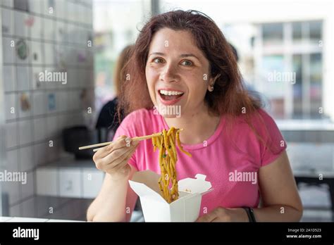 Portrait Of A Young Cheerful Girl Eating Chinese Noodles In A Cafe And Looking Out The Window