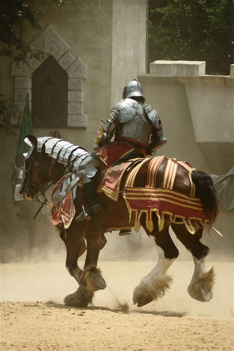 Two Men Dressed In Armor Riding Horses On Dirt Ground With Trees Behind