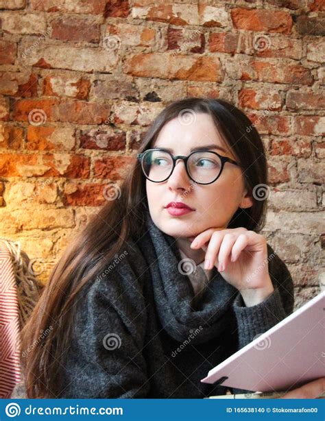 A Young Girl With Dark Long Hair And Glasses Sits On A Sofa In A Cafe