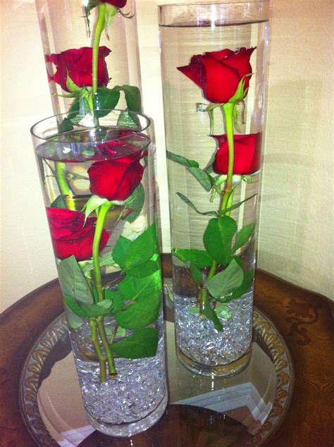 Two Clear Vases With Red Roses In Them On A Table Next To Each Other