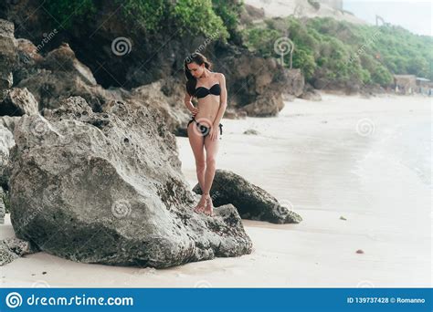 Tanned Brunette Girl In Black Swimwear Sunbathes On Beach With Rocks