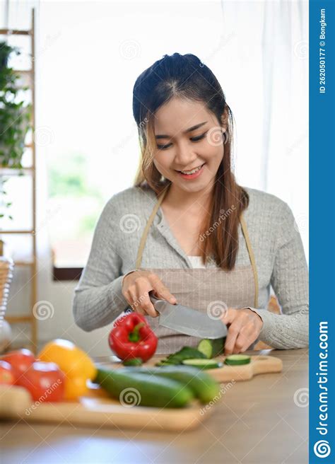 Beautiful Asian Female Housewife Wearing Apron Chopping Cucumber In The Kitchen Stock Image