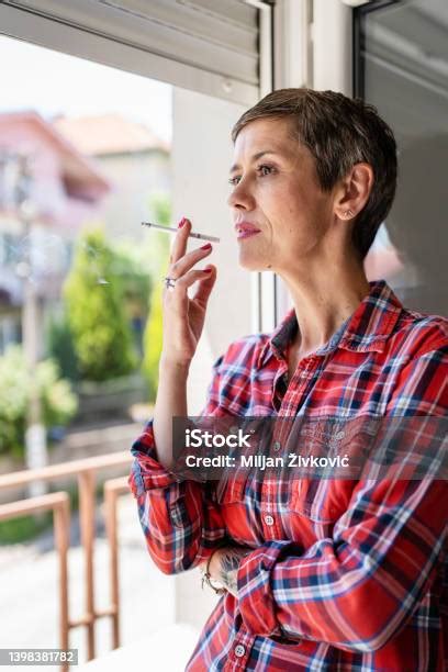 한 여성 성숙 또는 노인 여성 흡연 밝은 날에 집에서 창문으로 담배를 피우는 실제 사람들은 매일 공간을 복사합니다 건강에 해로운 습관 니코틴 중독 심각한 걱정을 기다리고있는