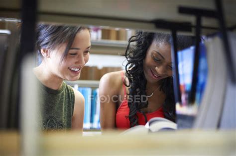 Two Young Female College Students Reading Book On Library Shelves