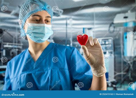 female doctor or nurse in blue uniform gloves mask holding small red heart in operating room
