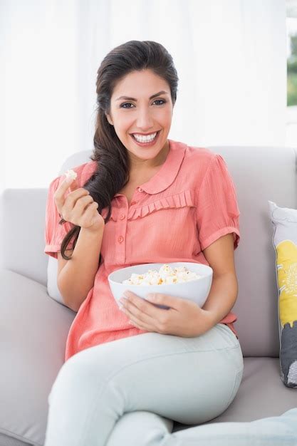 premium photo cheerful brunette sitting on her sofa eating bowl of popcorn