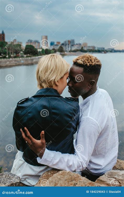 Interracial Couple Sits On Rocks And Hugs Against Background Of River