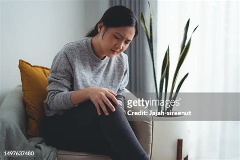 Asian Woman Suffering From Knee Pain Sitting On Armchair In Living Room