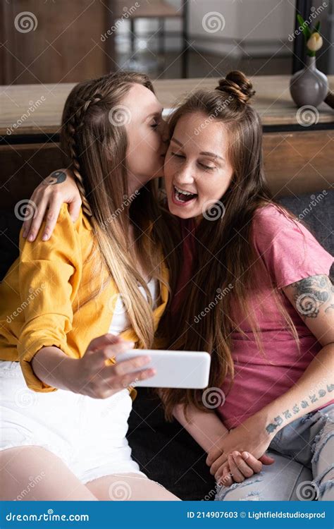 Lesbian In Yellow Blouse Kissing Girlfriend While Taking Selfie Stock