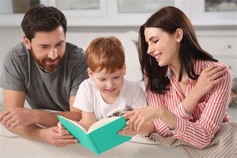 Happy Parents With Their Child Reading Book On Couch At Home Stock