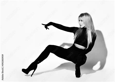 slim blonde woman with pouty lips in tight black overall suit high heeled shoes and gloves sits