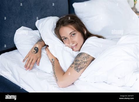 Attractive Tattooed Girl Lying In Bed And Looking At Camera In Bedroom