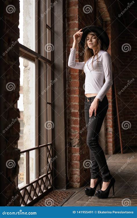 A Young Woman In A Hat Standing At The Window Posing For The Camera