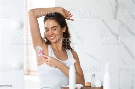 Young Woman Shaving Armpits Hair With Razor Standing In Bathroom Stock