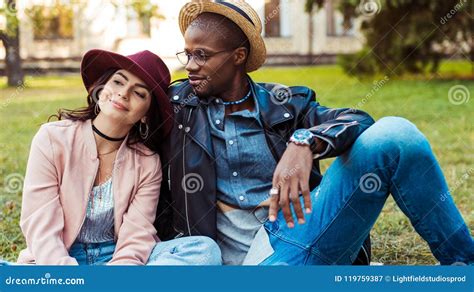 Pares Sonrientes Interraciales Felices En Los Sombreros Que Se Sientan En Una Hierba En Imagen