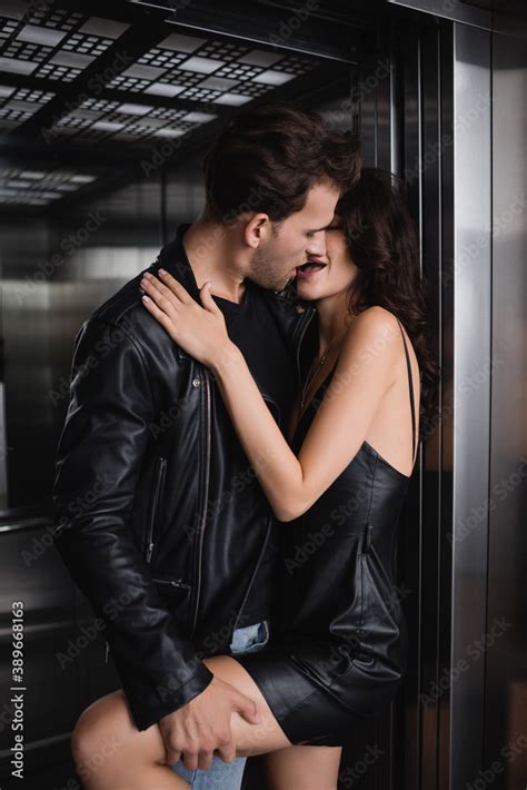 Man In Leather Jacket Kissing And Holding Leg Of Seductive Woman In Black Dress In Elevator