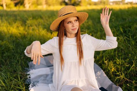 Portrait Of Displeased Red Haired Young Woman Wearing Straw Hat And White Dress Chasing Insects