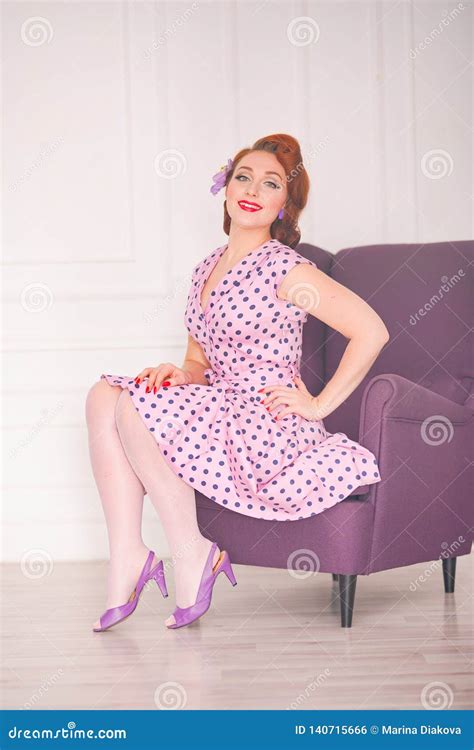Pretty Redheaded Pin Up Woman Wearing Pink Polka Dot Dress And Posing With Purple Armchair On