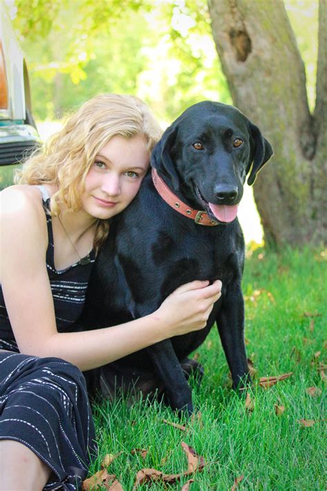 A Woman Is Sitting In The Grass With Her Black Dog Who Is Looking At