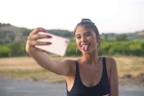 Portrait Of Teenage Girl Taking Selfie With Smartphone While Sticking Out Tongue Stock Photo