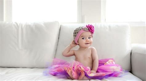 Cute Baby Girl Is Sitting On White Couch Wearing Colorful