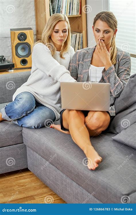 Two Young And Happy Women Are Sitting On A Couch With A Computer And Browsing Websites Stock