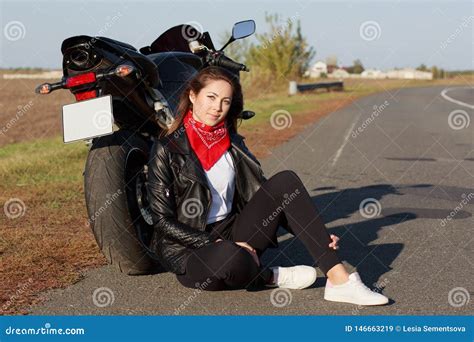 close up portrait of carefree female motorcyclist wears black clothes and white trainers sits